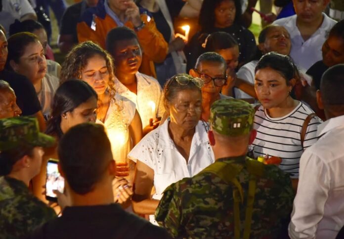 Una multitud de civiles con velas encendidas rodean a dos soldados uniformados en una velatón nocturna en Valledupar. El enfoque está en el rostro de una mujer mayor en el centro.
