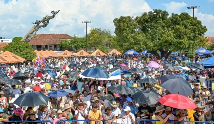 Multitud de turistas con sombreros y paraguas en la Plaza Alfonso López durante el **Festival Vallenato en Valledupar**, evidenciando el reto de movilidad.