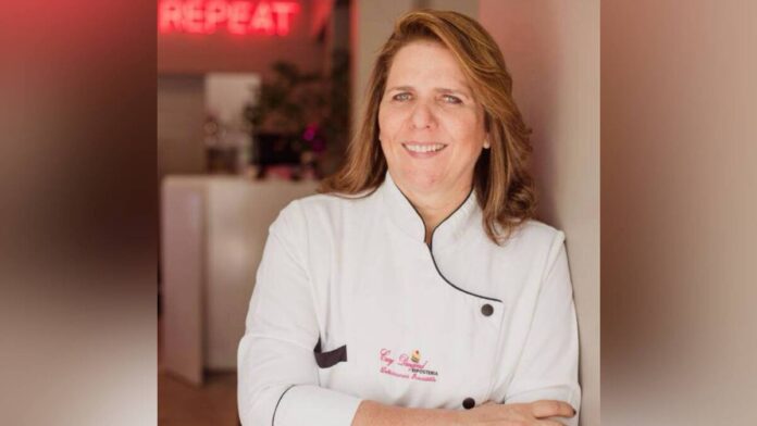 Retrato de la chef Cecy Dangond vistiendo su chaqueta blanca de cocina con el logo de su marca, sonriendo y apoyada en una pared.
