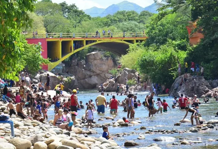 Multitud de bañistas disfrutan del río Guatapurí en el balneario de Hurtado en Valledupar, con el puente amarillo al fondo y montañas en la distancia.