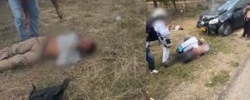 Escena de un accidente de tránsito en zona rural con personas heridas siendo auxiliadas junto a un automóvil negro chocado.