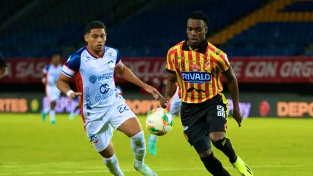 Dos jugadores de fútbol, uno de Alianza Valledupar con uniforme blanco y otro de Deportivo Pereira con uniforme amarillo y rojo, corren y disputan intensamente el balón en el campo de juego del estadio Hernán Ramírez Villegas durante un partido nocturno.