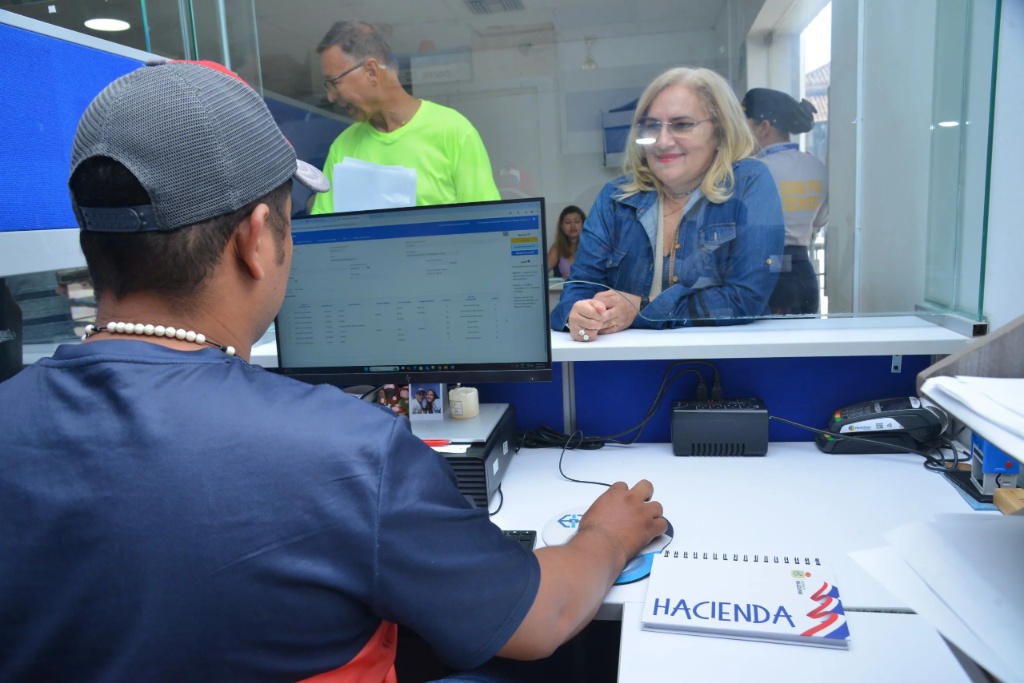 Un funcionario de la DIAN en Valledupar asesora a una mujer en ventanilla sobre cómo acceder a los alivios tributarios DIAN. En primer plano se observa un monitor y un cuaderno que dice "Hacienda".