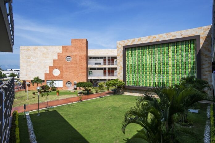 Vista panorámica a plena luz del día del moderno campus de la sede Areandina Valledupar. Se observa un edificio con una arquitectura distintiva que combina ladrillo marrón y una gran fachada de vidrio con un patrón verde vibrante, rodeado por un jardín interno bien mantenido con césped, palmeras y caminos de piedra.
