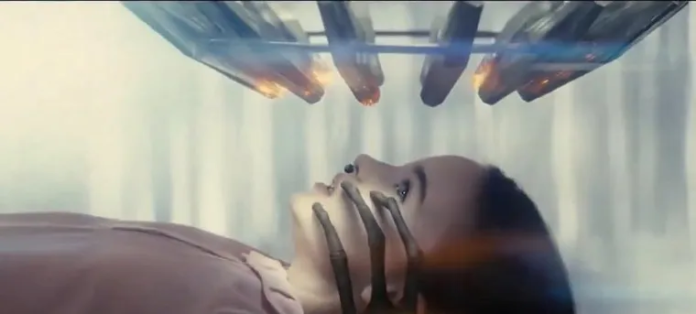 Escena de la película 'Disclosure Day' de Steven Spielberg, mostrando a una mujer mirando un dispositivo de examen con luces flameantes y una estructura mecánica cerca de su rostro, en un entorno de laboratorio de ciencia ficción oscuro.