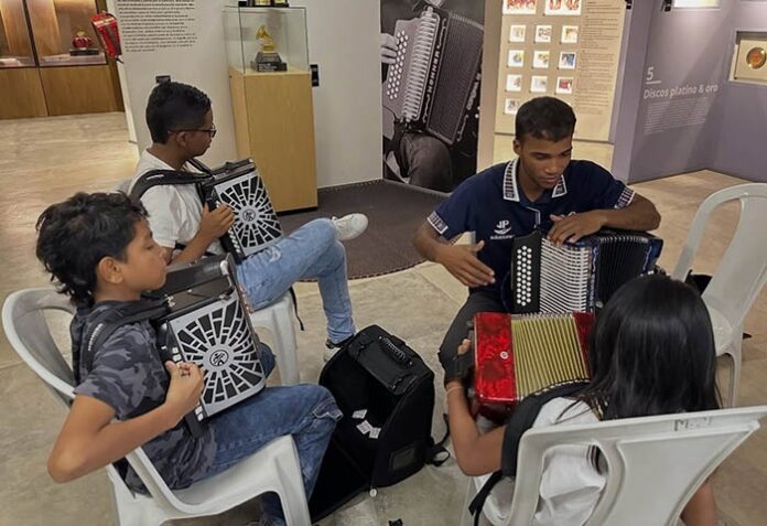 El rey vallenato Gonzalo Arturo ‘El Cocha’ Molina, vistiendo camisa blanca y gorra negra, de perfil, observa y orienta a un niño acordeonista con gafas que posa sonriente con su instrumento en el centro comercial Guatapurí, Valledupar. Representa el programa Huellas del Maestro de la Fundación Cocha Molina.