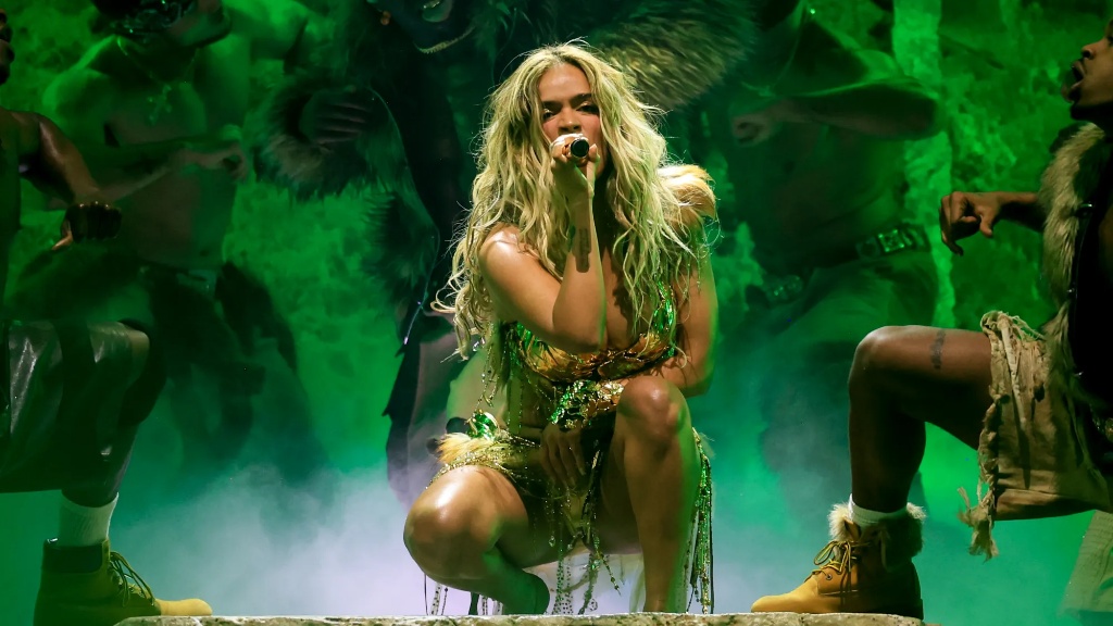 Karol G en Coachella 2026 cantando en cuclillas con un atuendo dorado brillante bajo una iluminación verde esmeralda y humo.