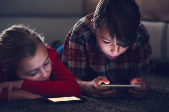 Dos niños acostados en una alfombra en la oscuridad, con la luz de las pantallas de celulares iluminando sus rostros, ilustrando riesgos de la falta de seguridad digital infantil.