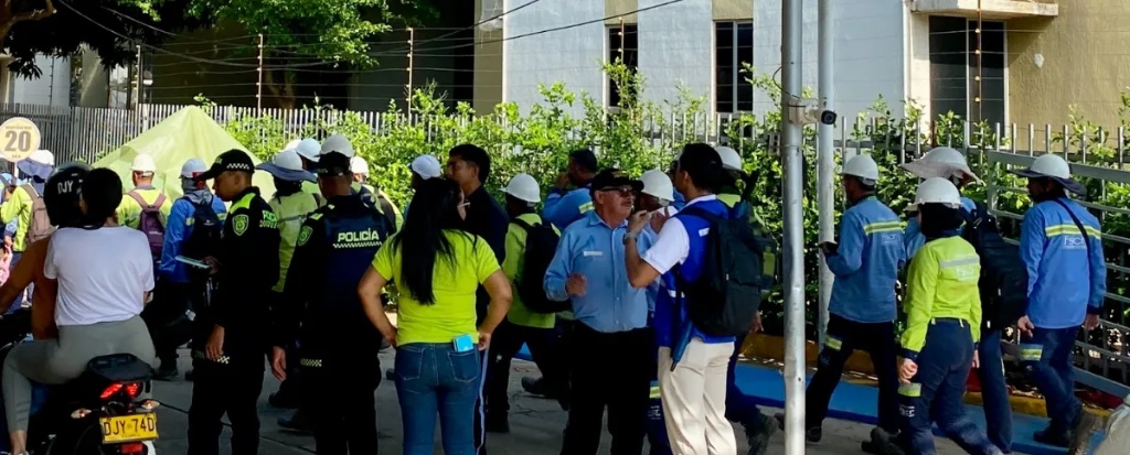 Un grupo de operarios de Afinia en Valledupar, vistiendo uniformes de color azul y verde con cascos blancos, se encuentra en una calle de la urbanización OGB junto a varios agentes de la Policía Nacional con uniformes verdes y una mujer con camiseta amarilla brillante. Se observa un ambiente de diálogo tenso y confrontación.