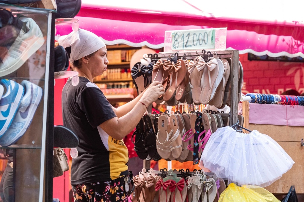Una mujer vendedora ambulante con turbante blanco y camiseta negra con amarillo arregla sandalias en un puesto callejero en Valledupar, bajo un toldo fucsia, representando la alta tasa de informalidad laboral descrita en la nota.